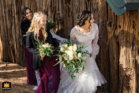 In South Lake Tahoe, California, a wedding photographer captures a shy and tender moment. The bride peeks from behind a rustic wood fence, her face expressing the quiet anticipation of her outdoor ceremony.